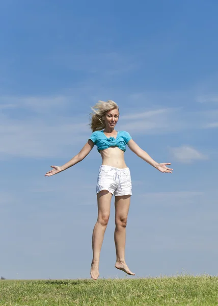 Счастливая женщина в белом бикини и шортах прыгает в летнем зеленом поле на фоне голубого неба — стоковое фото
