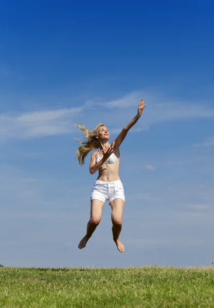 Счастливая женщина в белом бикини и шортах прыгает в летнем зеленом поле на фоне голубого неба — стоковое фото