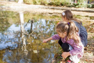 Two little girls playing in park near lake. Reflection of trees on water. Autumn sunny day.