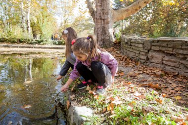 Two little girls playing in park near lake. Reflection of trees on water. Autumn sunny day.
