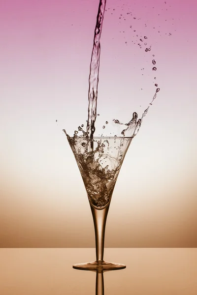 Wasser fällt in ein Glas und spritzt auf rosa und orangefarbenem Hintergrund — Stockfoto