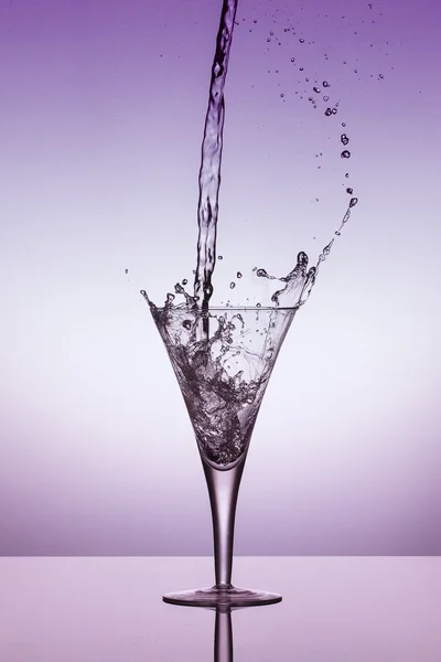 Вода потрапляє в склянку і бризкає на фіолетовому тлі — стокове фото