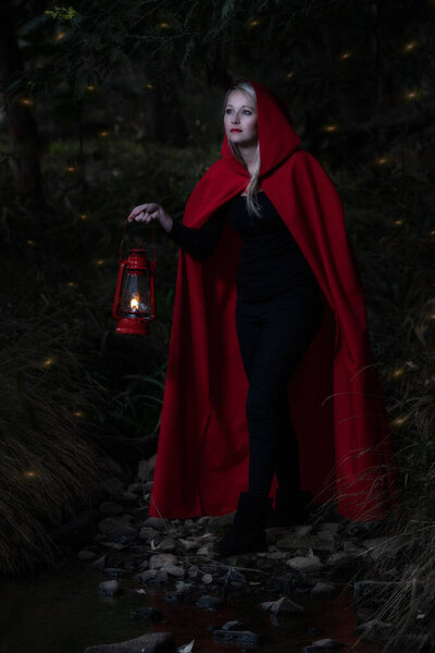 Attractive woman dressed a little red riding-hood cross a stream in a dark forest with a lantern