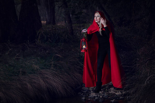 Attractive woman dressed a little red riding-hood cross a stream in a dark forest with a lantern