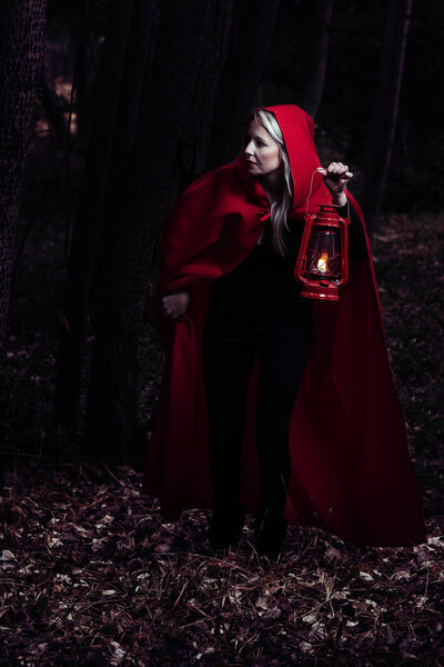 Attractive woman dressed a little red riding-hood walk in a dark forest with a lantern