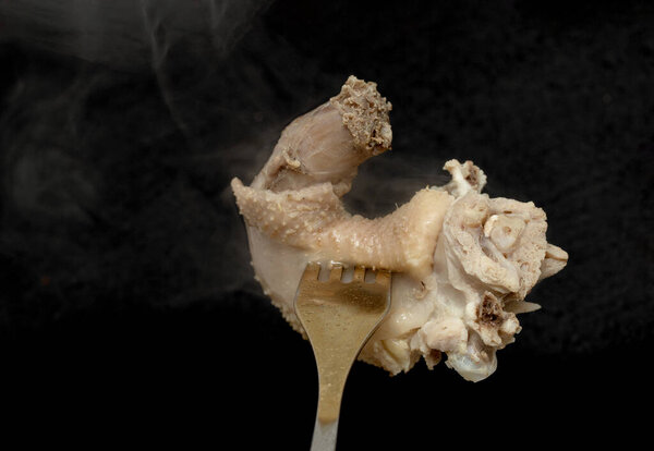 A piece of cooked chicken meat on a fork, steam smoke, black background.
