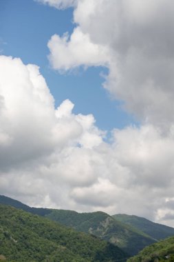 The sky is cloudy and the sun is shining through the clouds. The mountains are covered in trees and the sky is blue