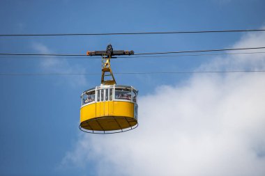 Havada sarı beyaz bir teleferik asılı duruyor. Yolcular gezinin ve manzaranın tadını çıkarıyorlar.