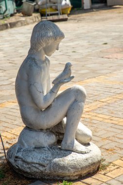 Kuşu tutan bir çocuğun heykeli. Heykel beyazdır ve pürüzlü bir dokusu vardır. Çocuk bir kayanın üzerinde oturuyor ve kuş onun eline tünemiş. Sahne huzur ve sükunet hissi veriyor.