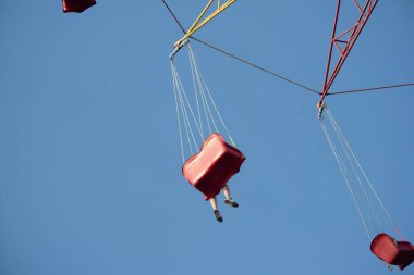 Bir insan havada kırmızı bir salıncakta sallanıyor. Gökyüzü mavi ve berrak. Salıncak kırmızı ve sarı bir ipten sarkıtılmış.