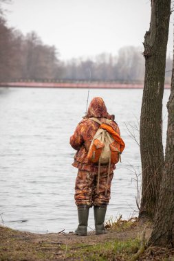 Kamuflaj kıyafeti giymiş bir adam gölde balık tutuyor. Su sakin ve gökyüzü kapalı.