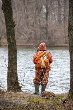 Kamuflaj kıyafeti giymiş bir adam nehir kıyısında elinde olta kamışı ile duruyor. Sahne sakin ve sakin, açık havanın ve suyun rahatlatıcı sesinin tadını çıkaran bir adam.