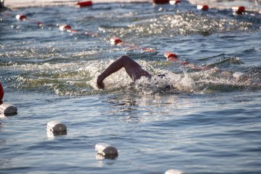 Suda bir yüzücü var ve arka planda başka yüzücüler de var. Su sakin ve berrak, ve tüm yüzücüler mayo giyiyor. Manzara huzurlu ve rahatlatıcı.