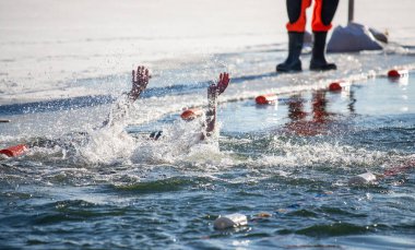 Bir adam suda yüzüyor ve başka bir adam onu izliyor. Sudaki adam kırmızı bir gömlek giyiyor.