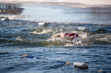 Suda bir yüzücü var ve yakınlarda bir kaç plastik şişe yüzüyor. Su soğuk ve dalgalı görünüyor, ve yüzücü kırmızı bir mayo giyiyor.