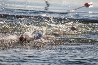 Bir adam elinde bir şişeyle suda yüzüyor. Su sıçrıyor ve adam kırmızı bir şapka takıyor.