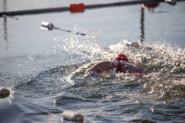 Suda bir yüzücü var ve su onun etrafına sıçrıyor. Sahne canlı ve enerjik, yüzücünün kırmızı mayosu ve suyun hareketi hareket ve heyecan hissi yaratıyor.