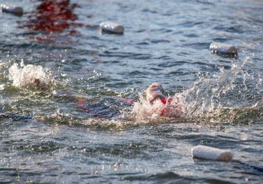 Kırmızı mayolu bir adam havuzda yüzüyor. Etrafına su sıçrıyor ve suda birkaç beyaz köpük şamandırası var.