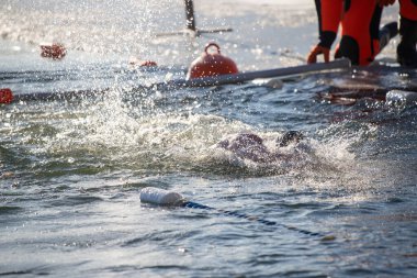 Suda bir adam yüzüyor ve bir cankurtaran onu izliyor. Cankurtaran turuncu bir takım elbise giyiyor.
