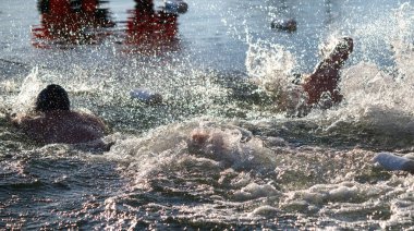 Bir grup insan, kırmızı gömlekli bir kişiyle gölde yüzüyorlar. Su dalgalı ve yüzücüler etrafa sıçrıyor.