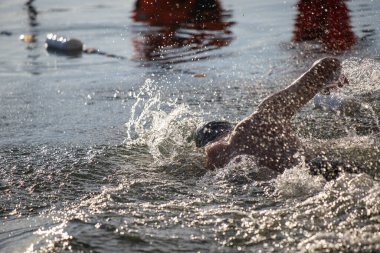 Bir yüzücü suyun içinde, etrafına su sıçratıyor. Su sakin ve berrak, ve yüzücü siyah bir mayo giyiyor.
