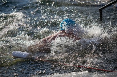 Bir adam elinde beyaz bir şişeyle okyanusta yüzüyor. Etrafına su sıçrıyor ve mavi bir şapka takıyor.
