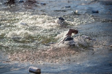 Suda bir grup yüzücü var, biri siyah gömlek giyiyor. Su bulanık ve etrafta plastik şişeler yüzüyor.