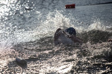 Bir adam elinde bir şişeyle suda yüzüyor. Su dalgalı ve adam etrafa sıçrıyor.