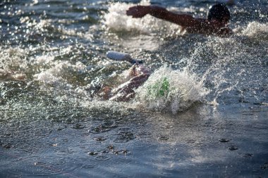 İki kişi suda yüzüyor, biri yeşil bir yüzme cihazı takıyor. Su dalgalı ve yüzücüler etrafa sıçrıyor.