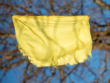 Sarı bir bez parçası çamaşır ipine asılı. Kumaş yırtılmış ve yıpranmış ve havada asılı duruyor. Arka plandaki mavi gökyüzü sahneye bir sükunet hissi katıyor