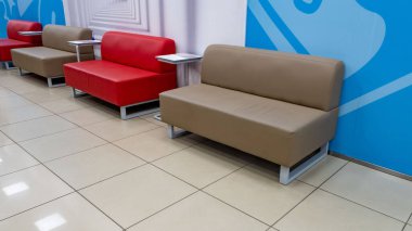 Arkalarında mavi bir duvar olan bir odada sıra sıra dizilmiş koltuklar. Kanepeler kırmızı ve kahverengi ve odanın ortasında beyaz bir masa var. Oda modern.