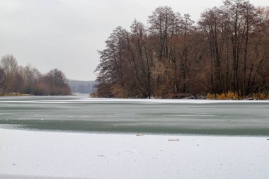 A serene winter scene with a frozen lake and trees in the background. The snow-covered landscape creates a peaceful and tranquil atmosphere. The frozen lake is surrounded by trees, which are bare