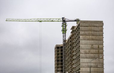A crane is lifting a large object over a building. The crane is green and yellow. The sky is cloudy and gray