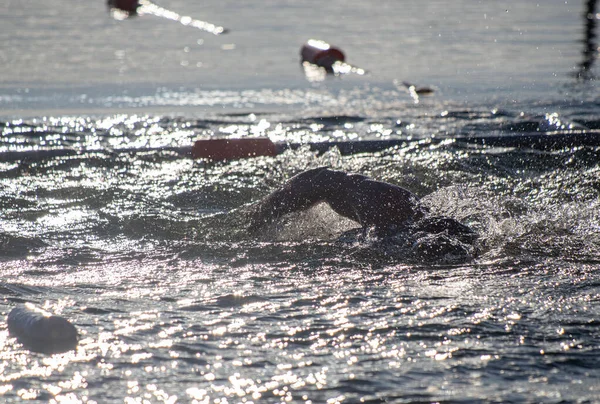 Suda bir yüzücü var ve arka planda birkaç kişi daha var. Su sakin ve güneş yüzeyde parlıyor.