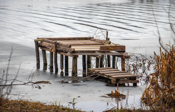 A wooden dock is floating in the water. The dock is old and has been abandoned. The water is calm and the dock is surrounded by weeds