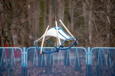 A blue and white object is hanging from a chain. The object is a chair. The chair is upside down and he is in a park. The scene is quiet and peaceful