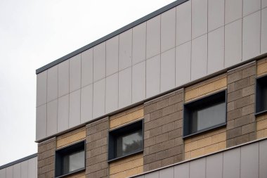 A building with a gray roof and tan bricks. The building has three windows on the side. The windows are rectangular and have a black frame. The building is tall and has a modern look