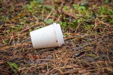 A cup is laying on the ground in a field. The cup is empty and has a lid on it. The field is covered in grass and leaves, and the cup is surrounded by dirt. The scene gives off a sense of abandonment