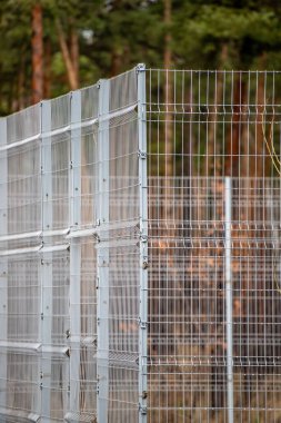 A fence with a reflection of the trees in it. The fence is made of metal and has a wire mesh