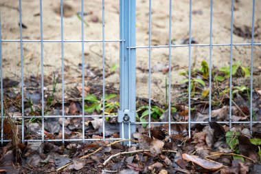 A metal fence with a gate is surrounded by a sandy area. The fence is covered in leaves and debris, giving it a somewhat unkempt appearance