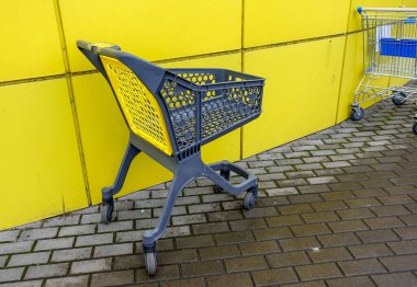 Kaldırımda sarı bir duvarın yanında bir alışveriş arabası oturuyor. Araba boş ve sarı bir sepeti var.