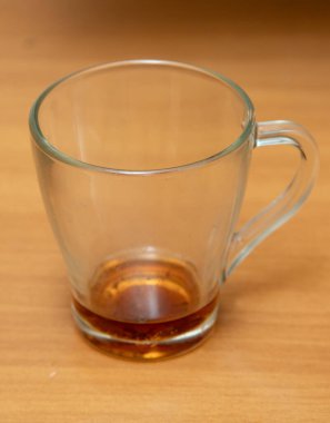 A glass cup is sitting on a wooden table. The cup is empty and has a brown liquid in it