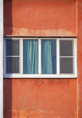 A window with a blue curtain is shown in front of a red wall. The window is open, and the curtain is drawn, allowing light to shine through