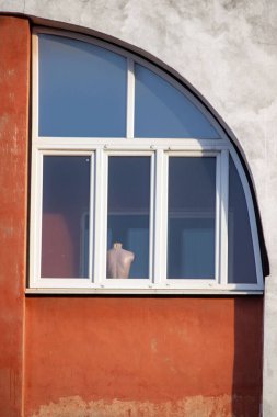 A man is standing in front of a window with a mannequin in the window. The mannequin is dressed in a white shirt and blue pants. The window is arched and has three panes of glass