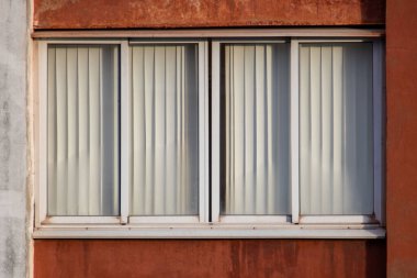A window with white curtains is shown in a building with a red wall. The curtains are drawn, and the window is open, letting in some light. Scene is calm and peaceful, as the curtains are drawn