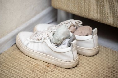 A pair of white shoes with a white sock inside. The sock is placed in the shoe, covering the sole. The shoes are placed on a rug, and the rug is located near a wall
