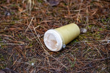 A plastic cup is on the ground in a field. The cup is empty and has a lid on it. The scene is desolate and abandoned, with no one around to clean up the trash