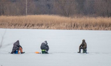 Üç adam donmuş bir gölde balık tutuyor. Adamlardan biri bankta oturuyor. Diğer ikisi suda oturuyor.