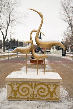 Üç kuş bir kaidede duruyor. Kuşlar altın ve beyaz. Heykel taştan yapılmıştır.