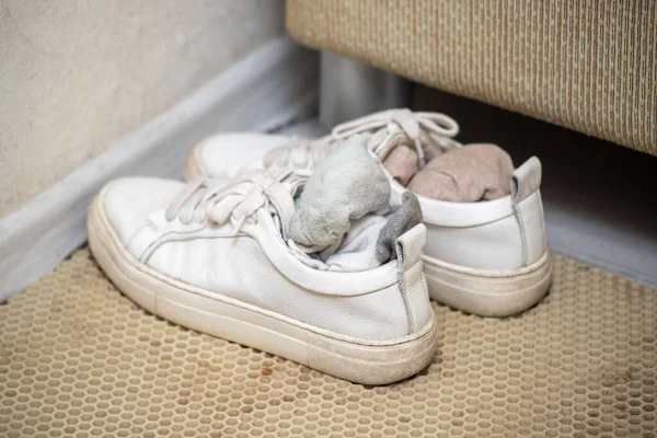 A pair of white shoes with a white sock inside. The sock is placed in the shoe, covering the sole. The shoes are placed on a rug, and the rug is located near a wall